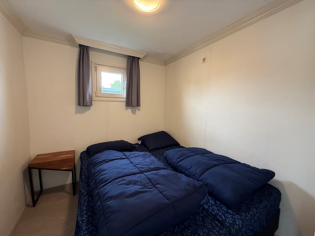 Slaapkamer met twee eenpersoonsbedden, houten nachttafeltje en een klein raam met gordijnen
