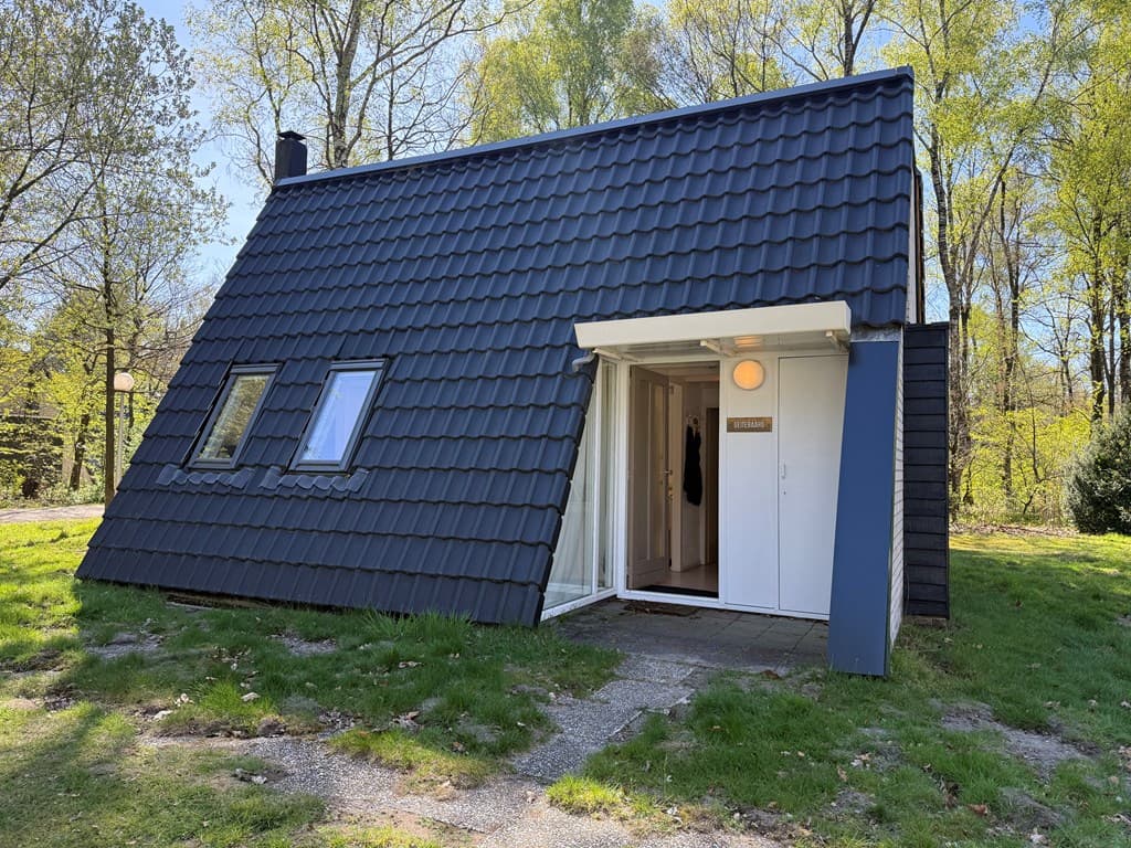 Zwart geglazuurde bungalow met schuine daken, een entree en groene omgeving met bomen en gazon.