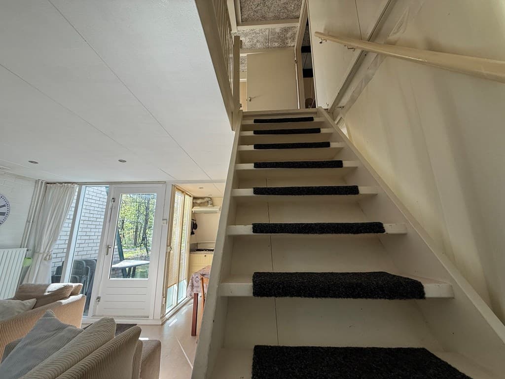 Een trap met tapijt naar de bovenverdieping in een bungalow, met zicht op een brede deur naar buiten.