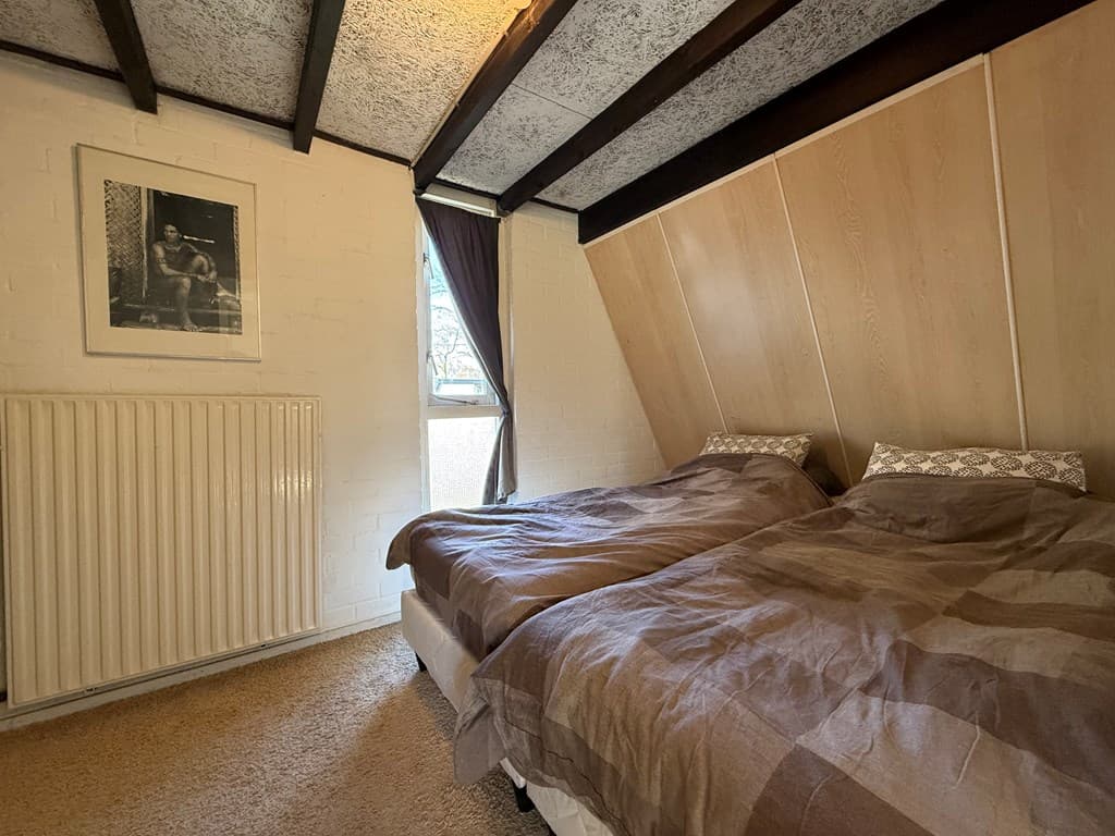 Slaapkamer met twee eenpersoonsbedden, houten plafondbalken en een raam met een gordijn, verder een radiator en een schilderi