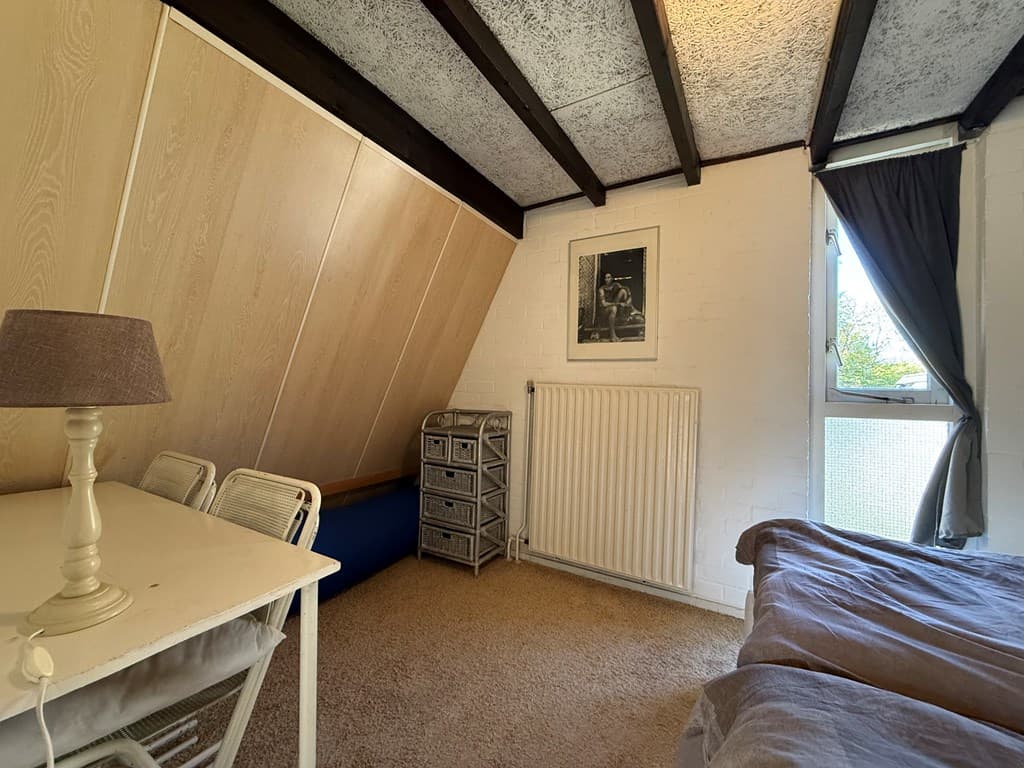 Slaapkamer met schuin dak, bed, bureau en stoel, radiator en uitzicht op buiten met verduisteringsgordijn.