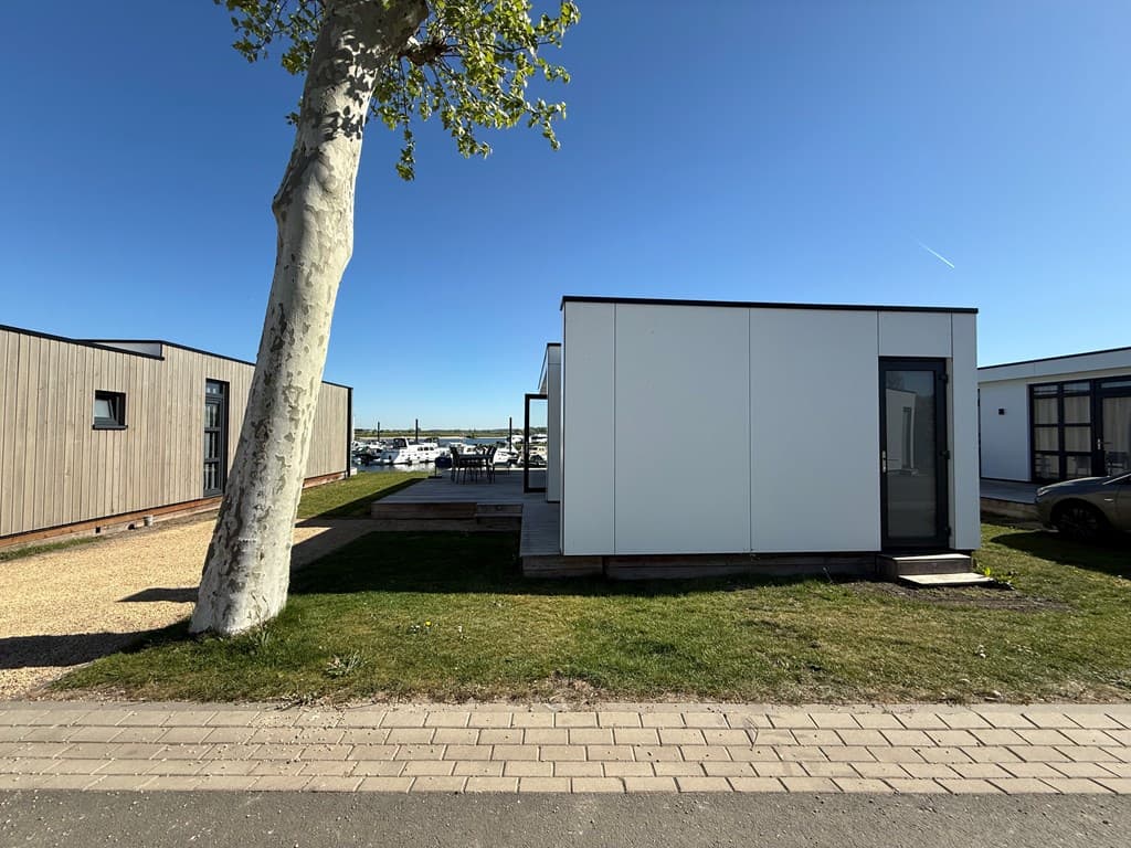 Moderne woning met witte gevel, grote ramen en een houten terras, gelegen bij een jachthaven onder een blauwe lucht.