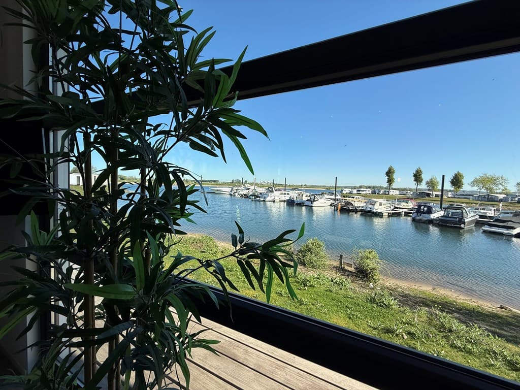 Uitzicht op de jachthaven met boten, gras en bomen aan de oever, zichtbaar vanuit een raam met een plant ernaast