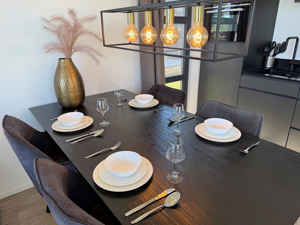 Eettafel met zes stoelen, wijze glazen, borden en een opvallende lamp erboven, een moderne keuken op de achtergrond