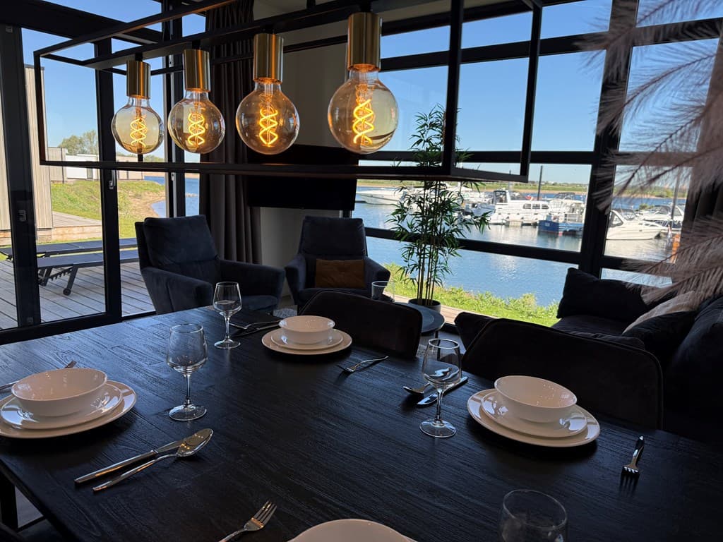 Eetruimte met uitzicht op het water, luxe stoelen en grote ramen met moderne verlichting boven de tafel.