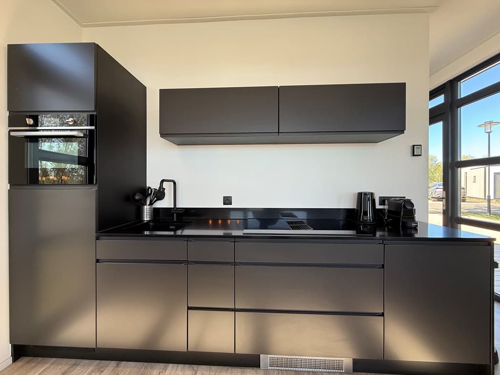 Moderne zwarte keuken met oven, koelkast en kookplaat, strak vormgegeven met minimalistische kastjes en verlichtingspaneel.
