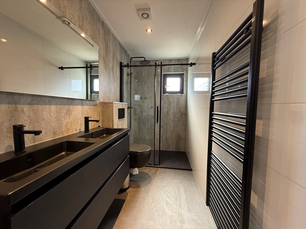 Moderne badkamer met zwarte wastafels, spiegel, douchecabine en handdoekradiator tegen een lichte tegelwand