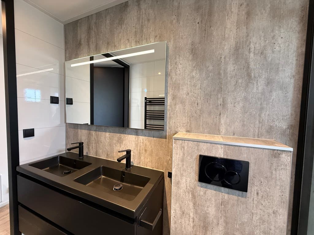 Moderne badkamer met dubbele wastafel, grote spiegel en wand met betonlook.