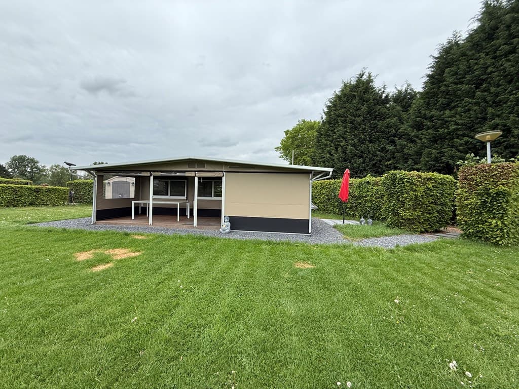 Vakantiehuis met een overdekt terras, omgeven door gras en hoge hagen, met een parasol rechts en een grindpad.