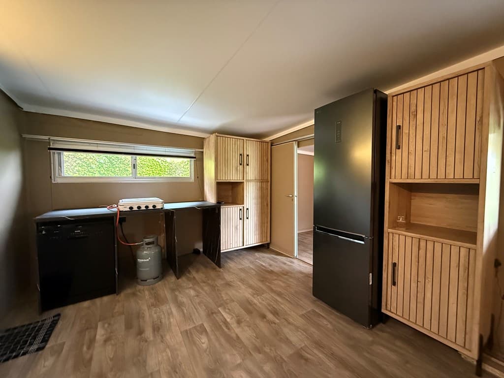 Keukenruimte met houten kasten, koelbox en gasfornuis, enkele ramen voor natuurlijke verlichting.