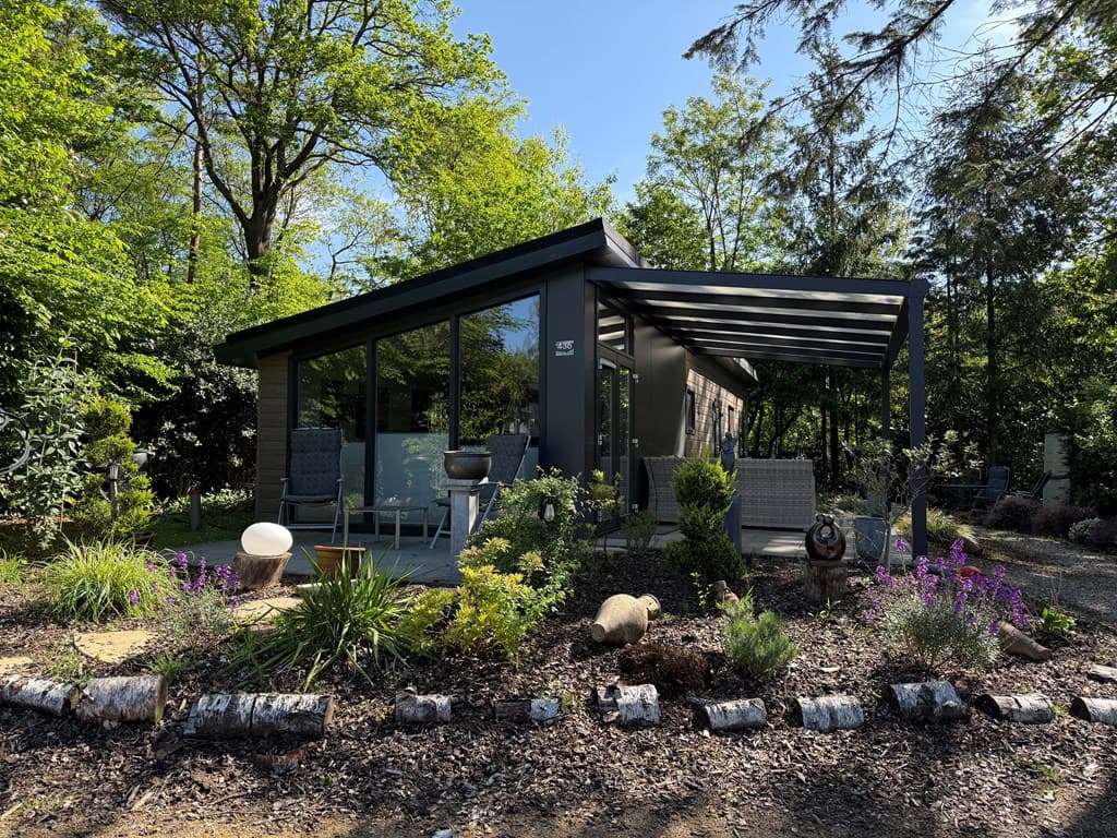 Moderne vakantiewoning met schuin dak en terras onder overkapping, omringd door bloeiende planten en groen.
