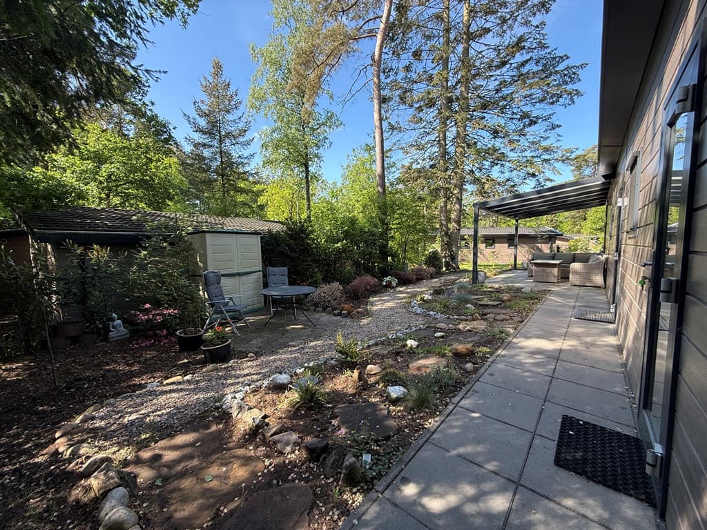 Tuin met grindpad, zitgedeelte en schuur, omgeven door bomen en planten bij de woning in Veluwepark de Bosgraaf.