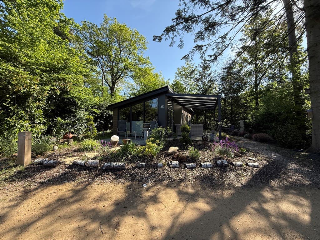 Moderne vakantiewoning omgeven door groen, met een oprit van zand en aangelegde tuin met bloemen en planten.