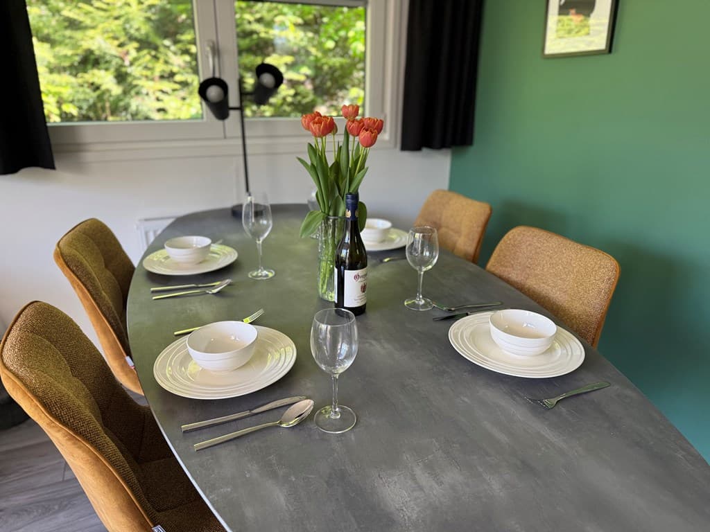 Eettafel met borden, glazen en een vaas met tulpen, omringd door stoelen in een groene kamer met rondom ramen