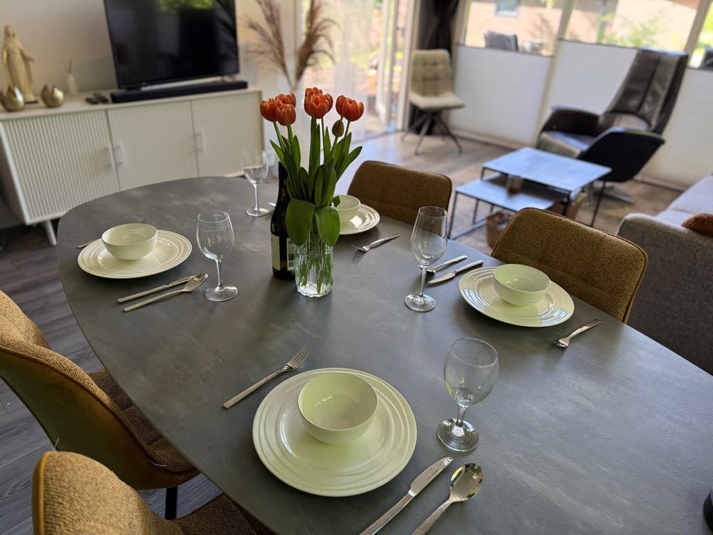 Eethoek met een grote tafel, vier glazen, borden en bestek, met een vaas bloemen op de tafel en een modern interieur.