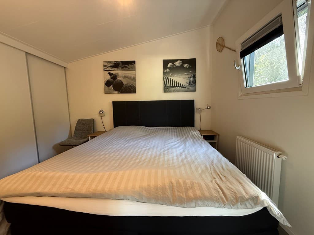 Slaapkamer met een groot bed, een bureau met stoel, en twee schilderijen aan de muur met een raam opzij.