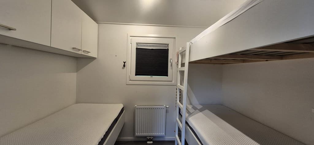 Slaapkamer met een stapelbed en een éénpersoonsbed, witte muren en een raam met jaloezieën.