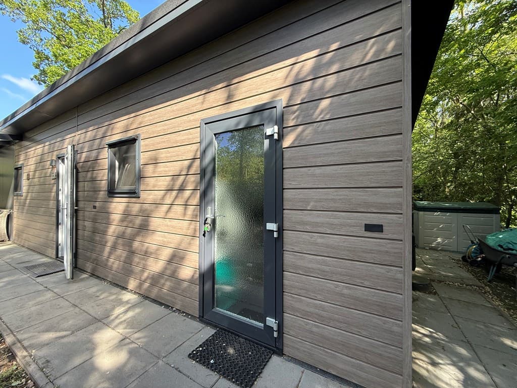 Moderne buitenzijde van een woning met een glazen voordeur, houten gevelbekleding en omliggende groene natuur
