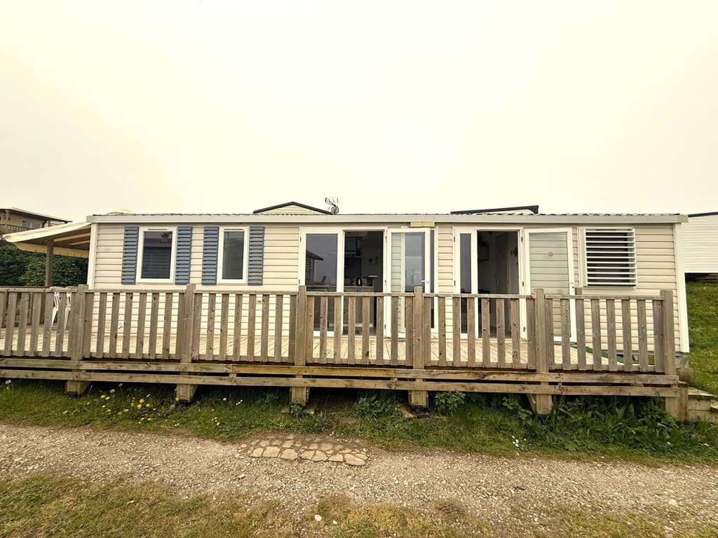 Vakantiehuis met houten terras en kunststof gevel, gelegen op een camping met groen grasveld en gravelpad eromheen
