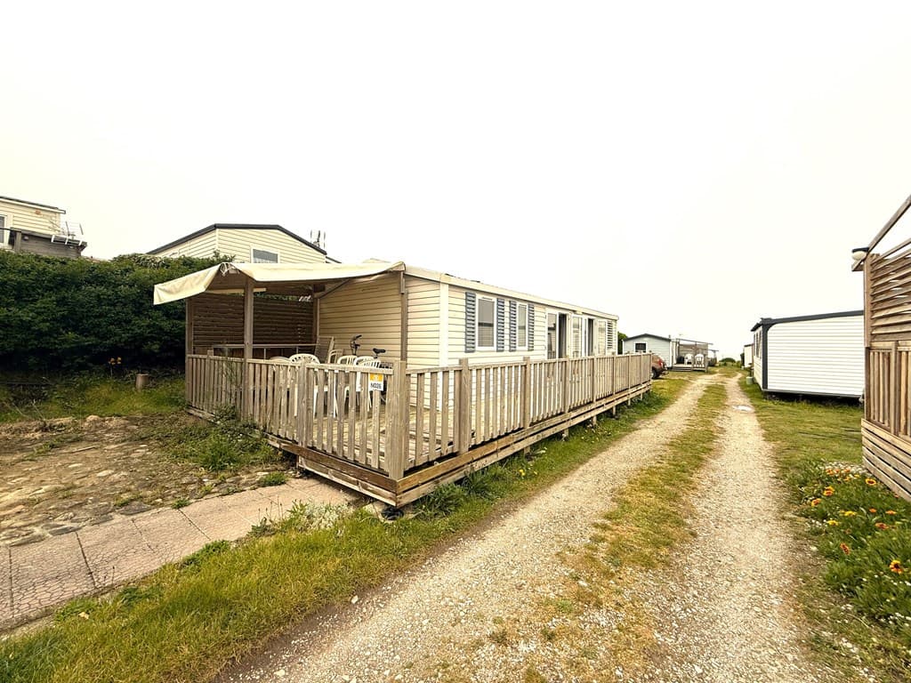 Mobiele woning met houten terras, gelegen aan een grindpad omringd door gras en andere woningen.