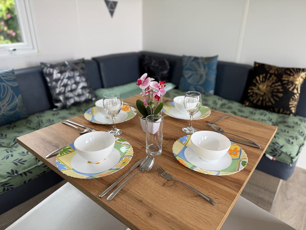 Eettafel met borden, glazen en bestek, omringd door een zithoek met kussens en een bloeiende plant in het midden