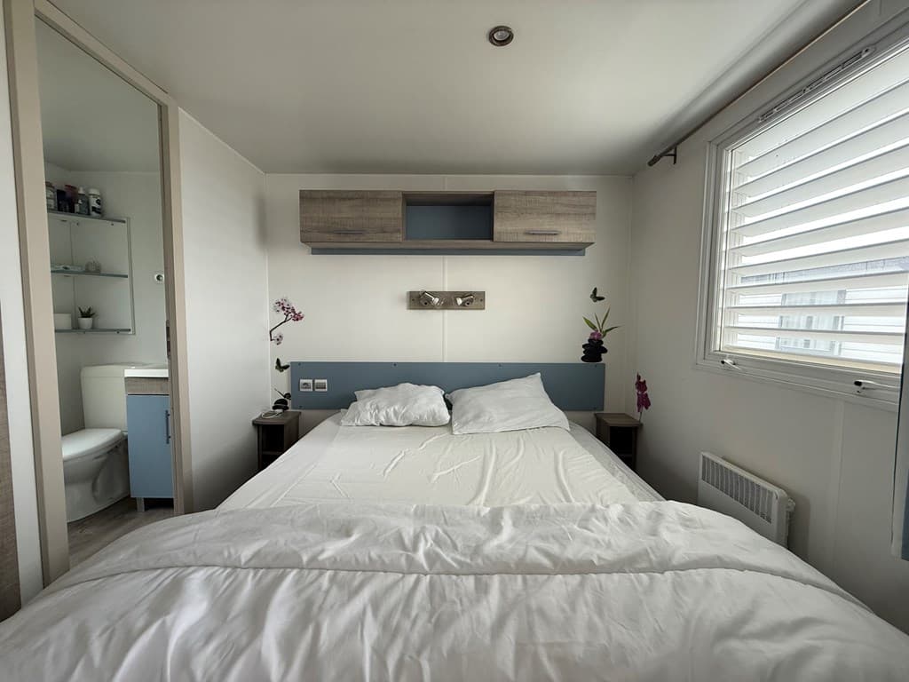 Slaapkamer met tweepersoonsbed, kastenwand en en-suite badkamer, lichte muren en raam met lamellen.