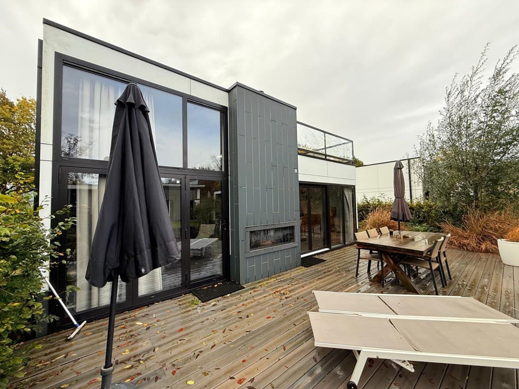 Moderne vakantiewoning met grote ramen, houten terras, ligstoelen en parasols, omringd door groen.