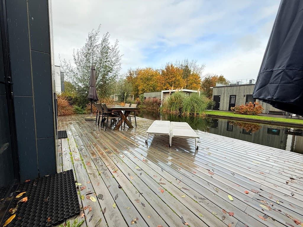 Terras met houten vlonders, tuinset en ligstoelen naast een vijver en omringd door bomen en struiken in herfstkleuren.
