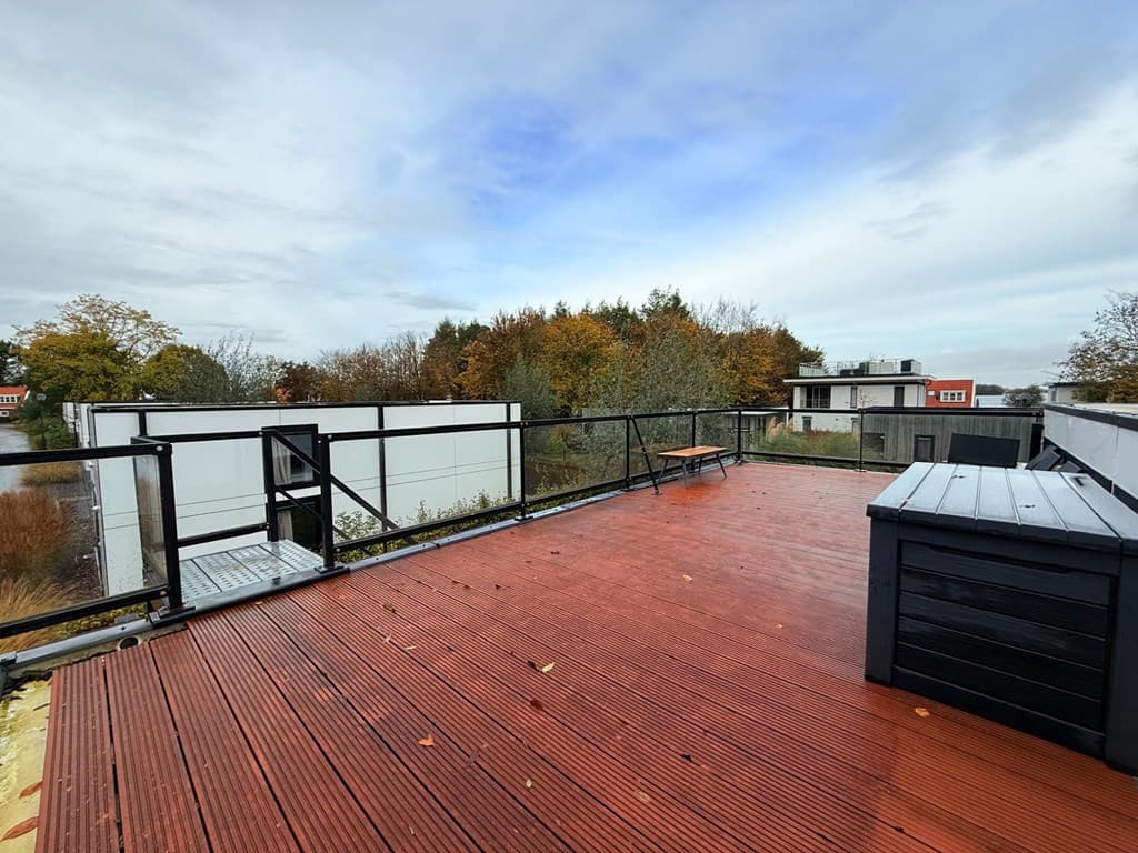 Terras met houten vloer en railing, uitzicht op natuur en omliggende woningen. Bewolkte lucht en herfstkleuren zichtbaar.