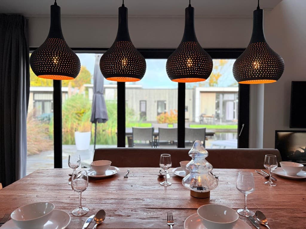 Eetkamer met houten tafel, vier zwarte lampen, uitzicht op terras met meubels en groene omgeving.