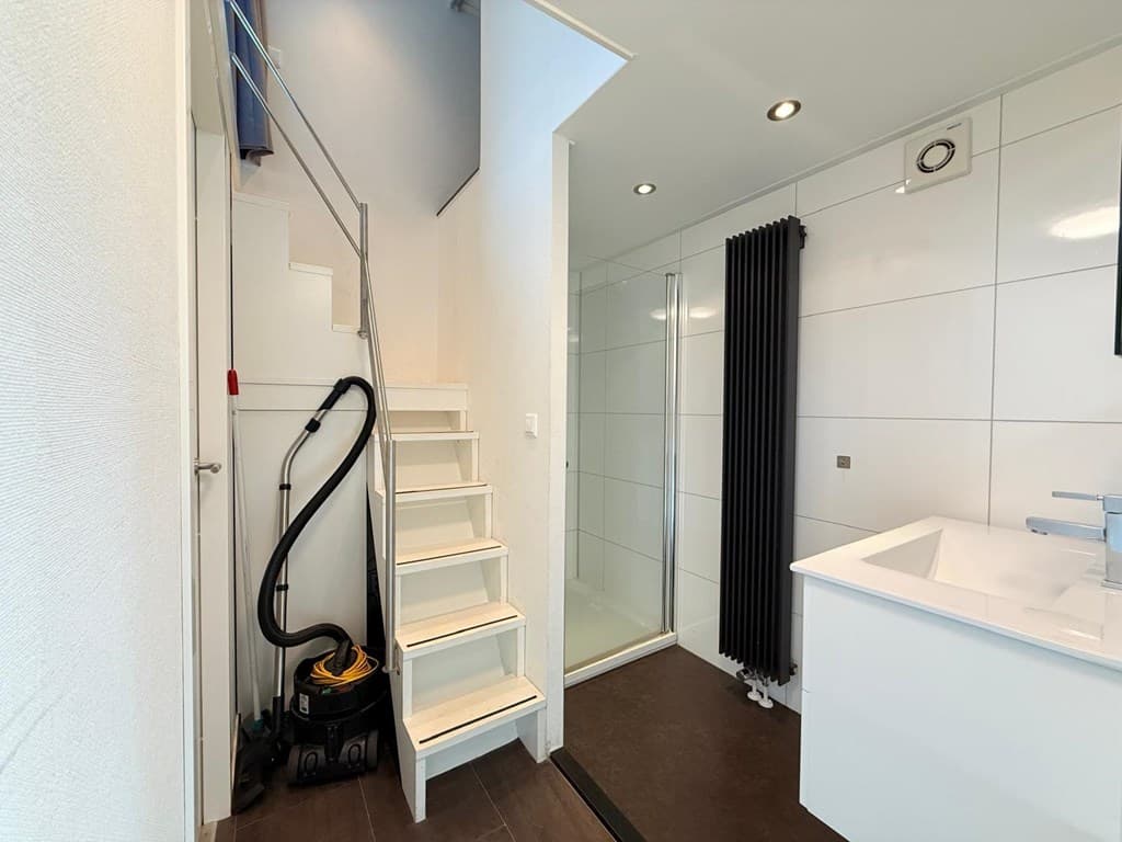 Entree met trap naar de bovenverdieping, toegang tot badkamer met douche, zwarte radiator en wastafel.