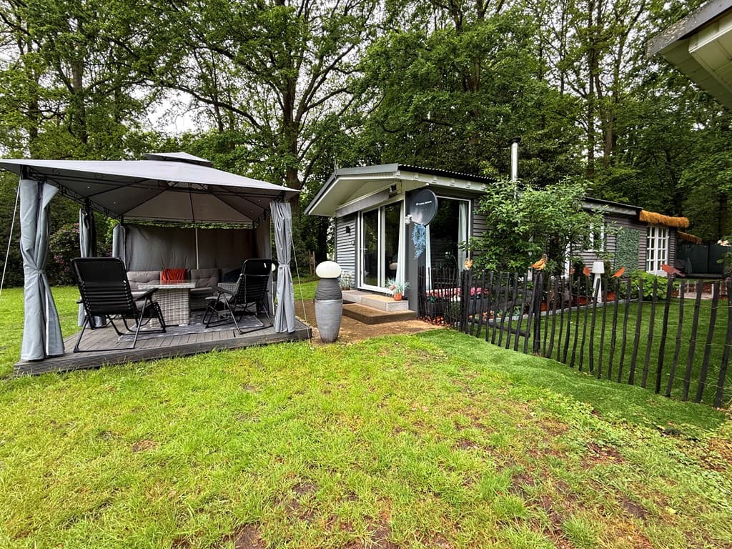 moderne vakantiewoning met een terrasoverkapping en groen omringd door bomen in een tuin met een hek