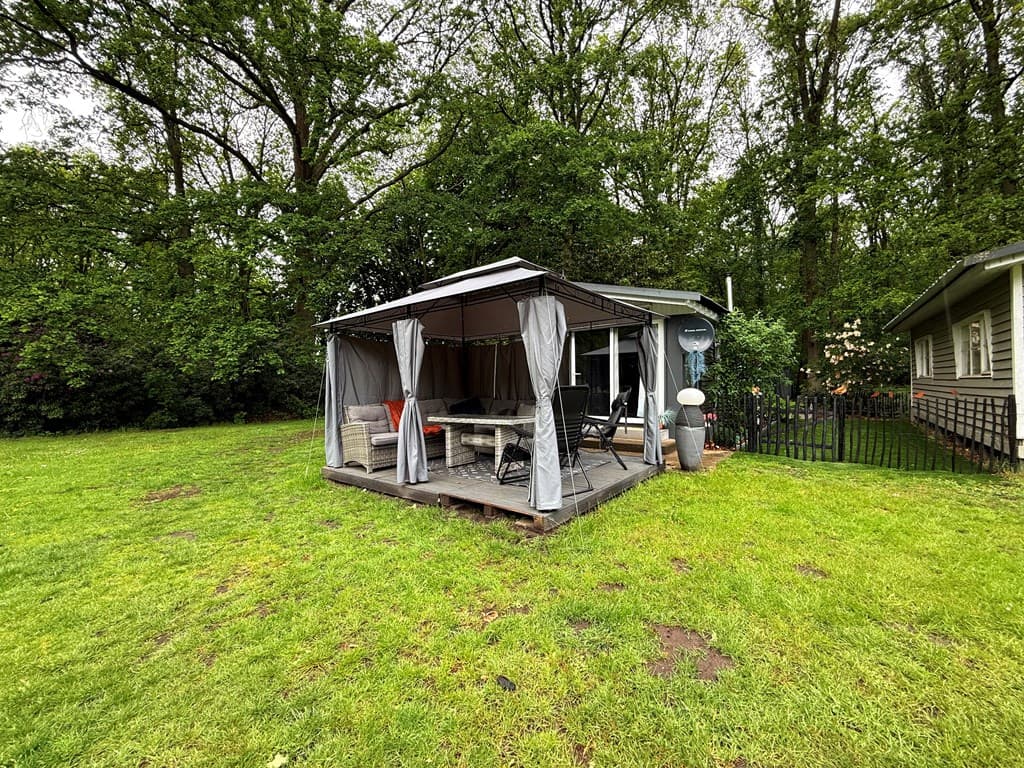 Tuinhuisje met overdekte zithoek op een houten terras, omringd door gras en bomen.