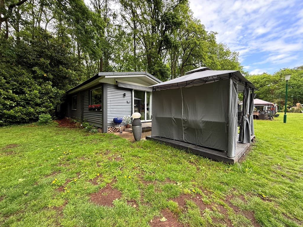 Zijkant van een woning met grijze houten gevel en terrasoverkapping in een groene omgeving met bomen en gazon.