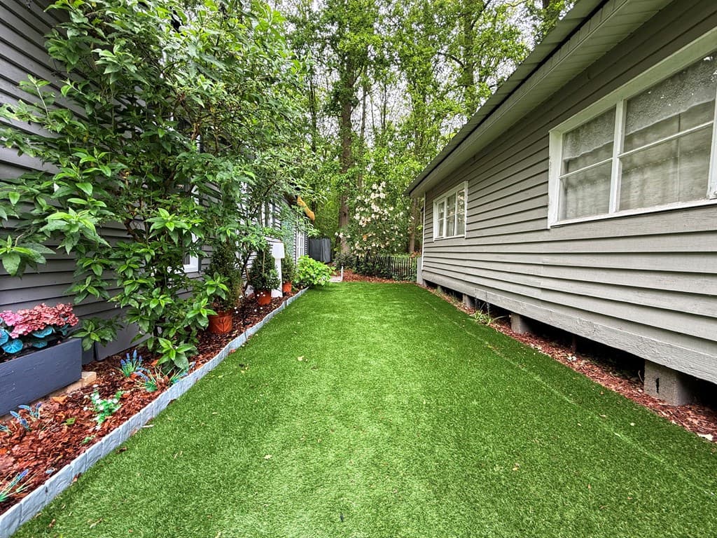 Groene tuin met kunstgras tussen twee woningen, omringd door bomen en struiken.