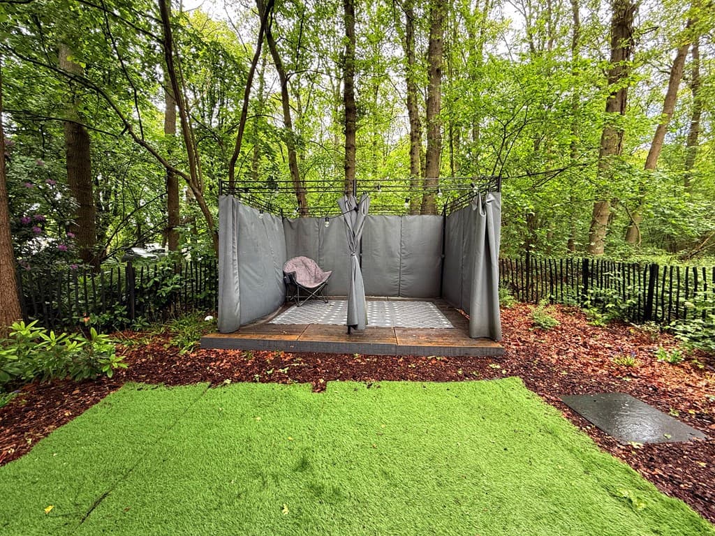 Tuin met groene grasmat, een betegeld terras en een afgeschermde zithoek tussen bomen.