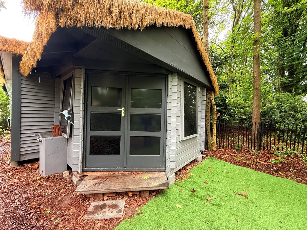 Houten bungalow met rieten dak, grote ramen en groene kunstgrassfeer rondom de ingang.