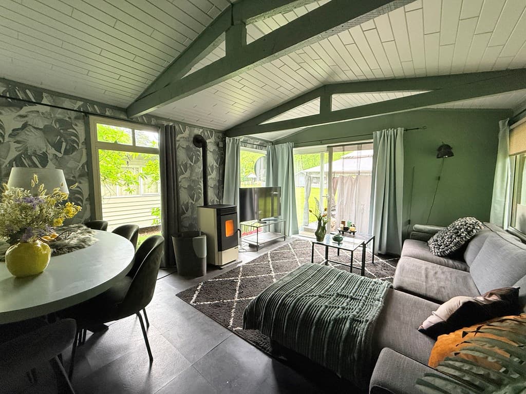 Woonkamer met groene muren, grote ramen, zithoek met bank en TV, en een ronde eettafel met stoelen.