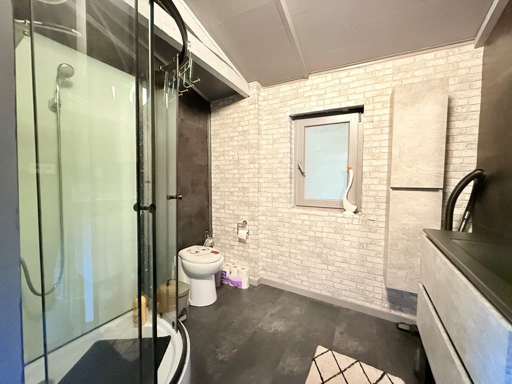 Badkamer met douche, toilet en raam, voorzien van lichte stenen muren en donkergrijze vloeren.