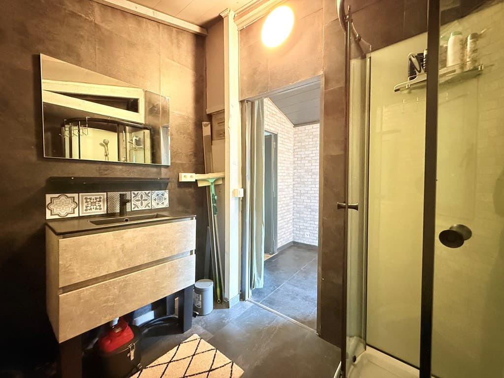 Badkamer met een douche, wastafelmeubel, grote spiegel en toegang tot een lichte ruimte met stenen muur.