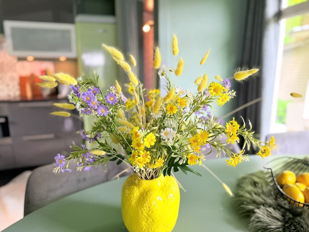 Een gele vaas gevuld met kleurrijke bloemen staat op een tafel in een moderne woonkamer met groene muren.