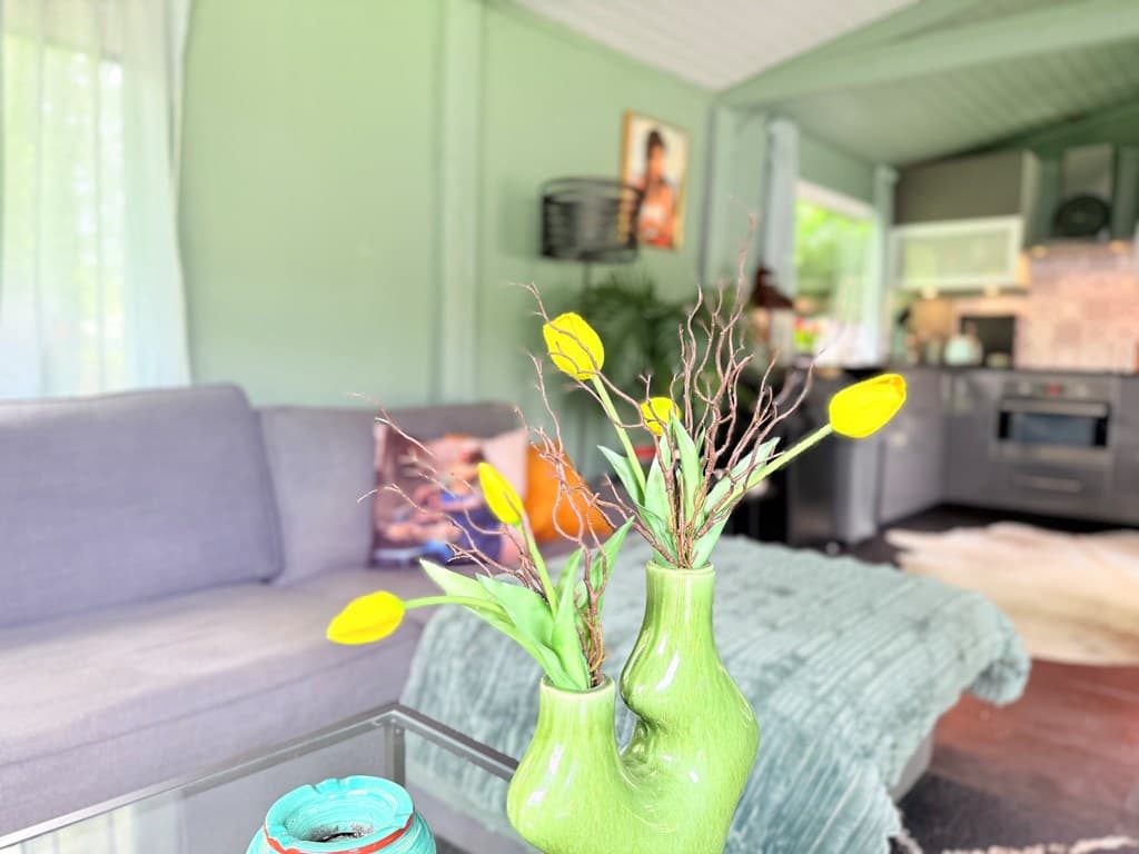 Ruime woonkamer met groene muren, een grijze bank en een vaas met gele bloemen op een glazen tafel.