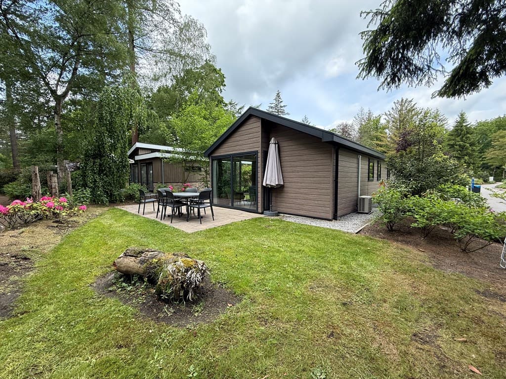 Moderne vakantiewoning met grote ramen, houten gevel, terras met tafel en stoelen, groene omgeving en bloemen.