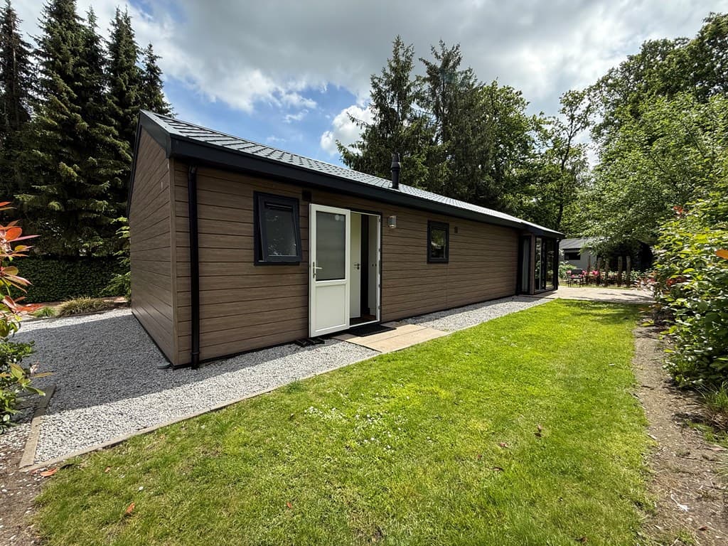 Moderne vakantiewoning met houten gevel, gras- en grindpad, omgeven door bomen en struiken in een groene omgeving.