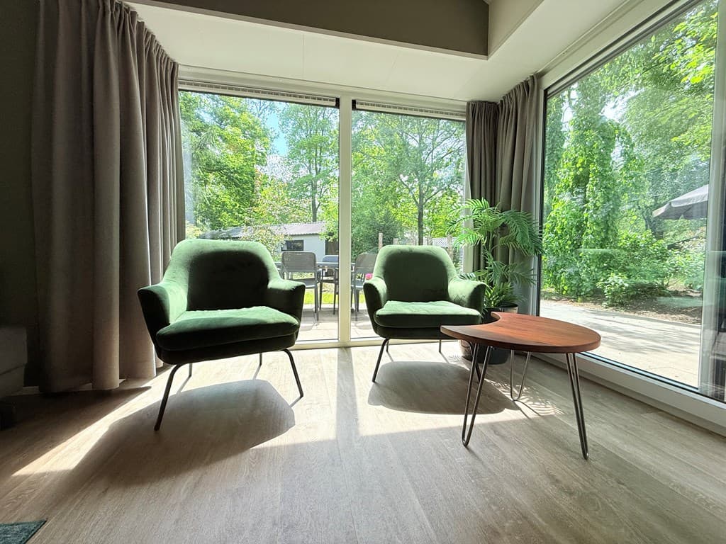 Twee groene stoelen en een ronde tafel bij grote ramen met uitzicht op buiten en bomen.
