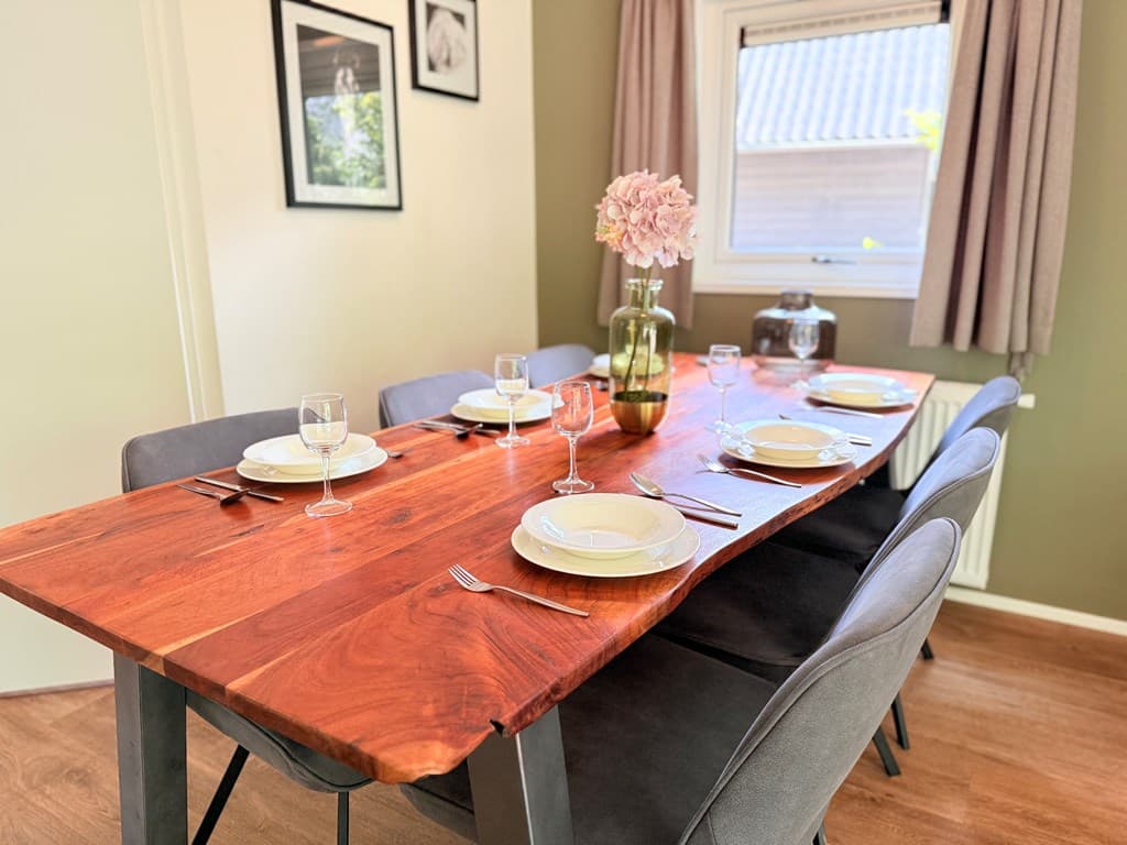 Eettafel met stoelen, gedekt met servies en glazen, in een sfeervolle eetkamer met groenachtige muren en ramen.