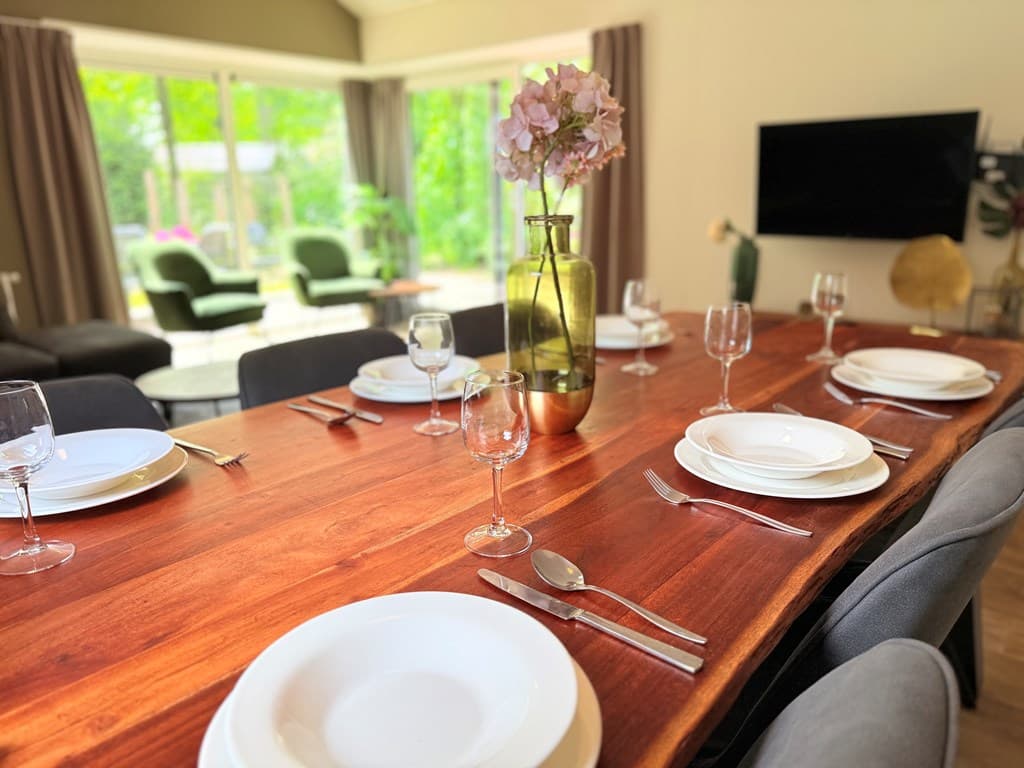 Eettafel met servies en glazen, zithoek op de achtergrond met groene stoelen en grote ramen naar de tuin.