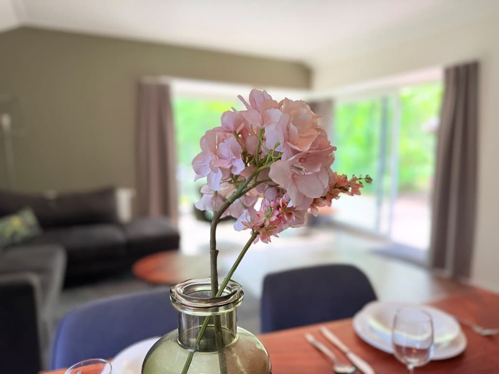 Bosachtige omgeving met een eetruimte, een vaas met roze bloemen en een zithoek op de achtergrond.