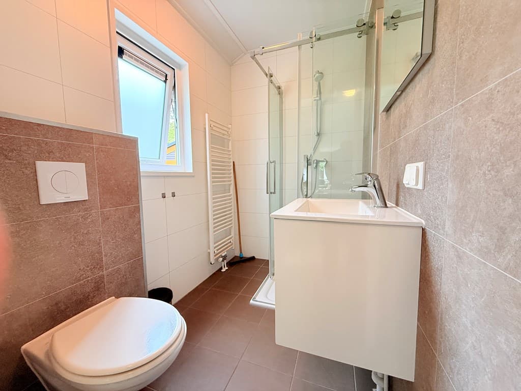 Badkamer met douche, toilet en wastafel, lichte tegels en venster voor natuurlijk licht