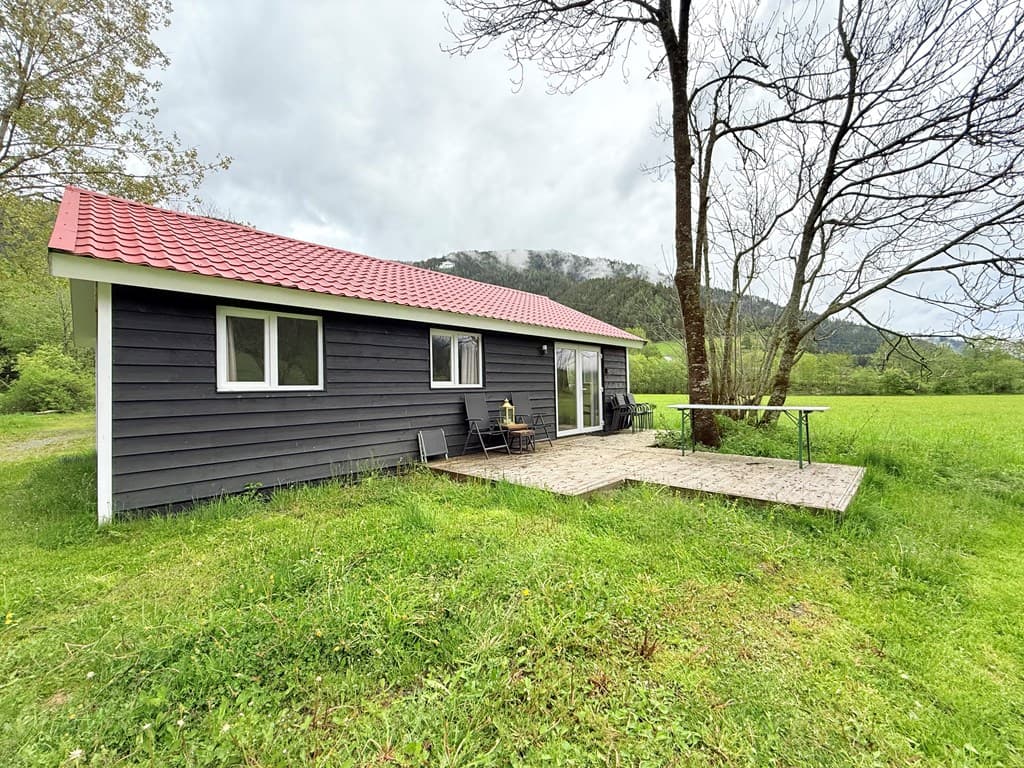 Zwart houten huis met rode dak, terras met tafeltje en stoelen, omgeven door groen gras en bomen met bergen op de achtergrond